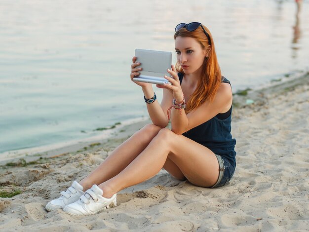 Mulher ruiva em shorts jeans e camiseta azul, sentado em uma praia e segurando o computador tablet.