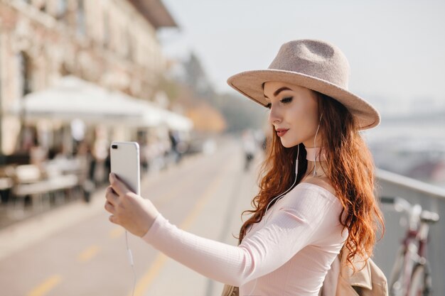 Mulher ruiva encantadora com smartphone fazendo selfie durante uma caminhada na cidade