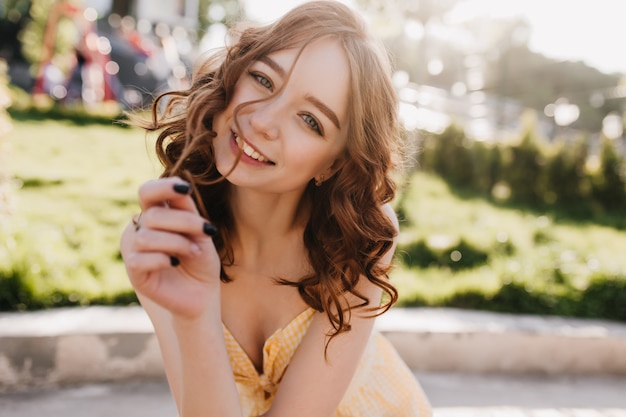 Mulher ruiva encaracolada interessada, sorrindo na manhã ensolarada. Adorável modelo feminino de gengibre, passando o dia de verão no parque.