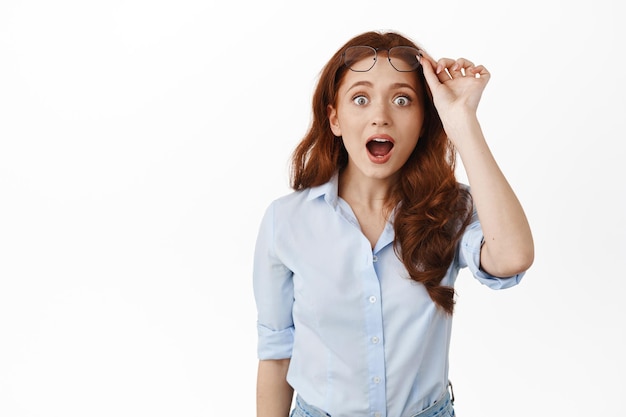 Foto grátis mulher ruiva fascinada e surpresa tira os óculos, deixa cair o queixo e diz uau impressionado, conferindo anúncio incrível, olhando para o banner com espanto, fundo branco