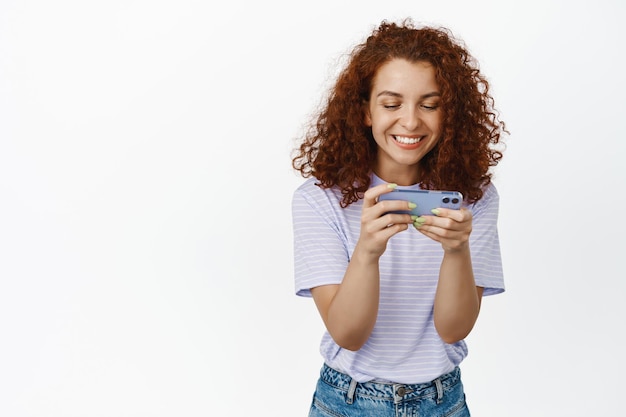 Mulher ruiva feliz sorrindo, jogando videogame no celular, segurando o smartphone horizontalmente, assistindo smth na internet celular, fundo branco