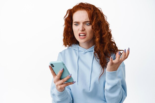 Mulher ruiva irritada e irritada fazendo uma careta de raiva, olhando para a tela do smartphone, lendo mensagem irritante, parada incomodada no branco