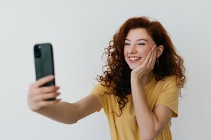 Mulher ruiva linda com cabelo encaracolado tocando seu penteado sorrindo e tirando selfie fazendo foto no smartphone em fundo branco