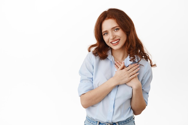 Mulher ruiva macia sorridente na blusa, segurando as mãos no coração e olhando romântico para a câmera, mantenha memórias reconfortantes na alma, de pé contra um fundo branco.