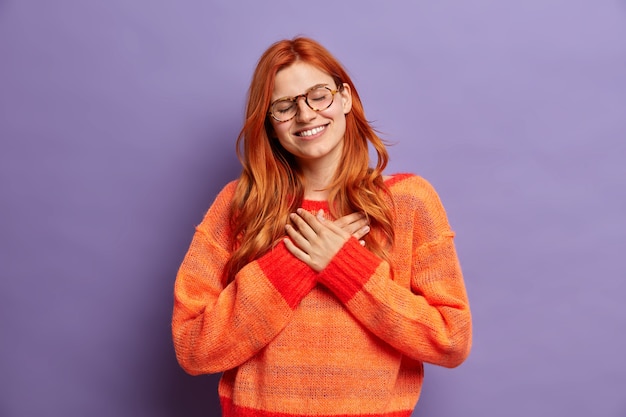 Mulher ruiva muito agradecida em óculos pressiona a mão no peito, fecha os olhos e sorri amplamente ouve palavras agradáveis vestida com um suéter enorme.