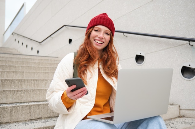 Mulher ruiva sorridente com telefone celular e laptop sentado na escada do lado de fora do prédio se conecta a pu