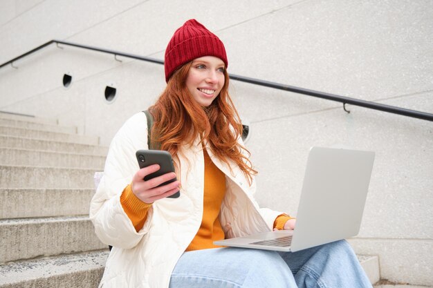 Mulher ruiva sorridente com telefone celular e laptop sentado na escada do lado de fora do prédio se conecta a pu