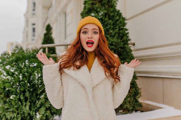 Mulher ruiva surpreendida de casaco branco acenando com as mãos na rua foto ao ar livre de uma garota espantada de casaco e chapéu
