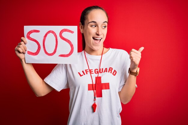 Mulher salva-vidas vestindo camiseta com cruz vermelha e apito segurando banner com mensagem sos apontando e mostrando com o polegar para o lado com cara feliz sorrindo