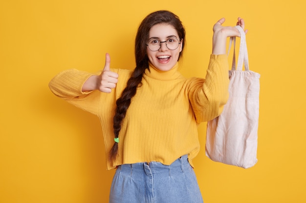 Mulher satisfeita com trança longa aparecendo polegar e segurando o saco na mão, desfrutando de suas compras