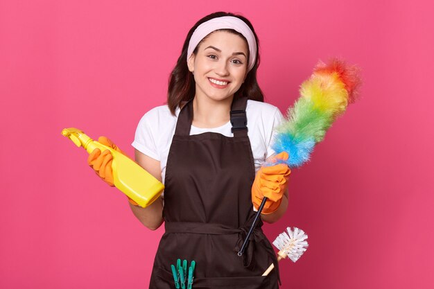 Mulher satisfeita vestida com camiseta casual, avental e faixa de cabelo, segurando detergente e espanador