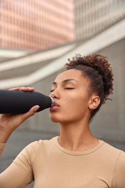 Foto grátis mulher saudável e em forma com cabelo encaracolado bebe água após um treinamento físico intensivo ao ar livre, se refresca com uma bebida energética e posa ao ar livre