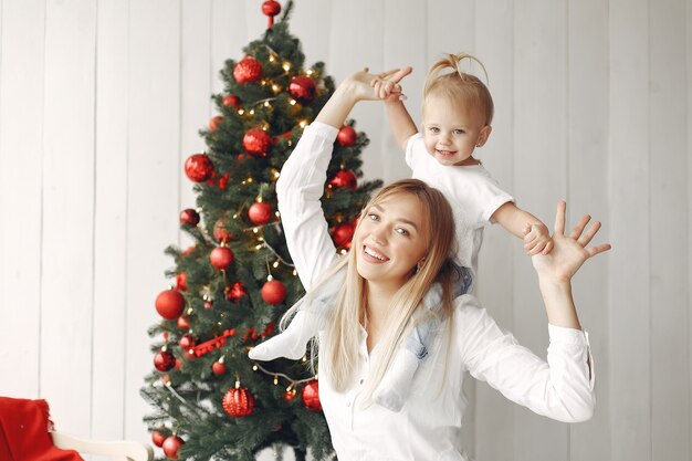 Mulher se diverte se preparando para o Natal. Mãe em uma camisa branca está brincando com sua filha. Família está descansando em uma sala festiva.