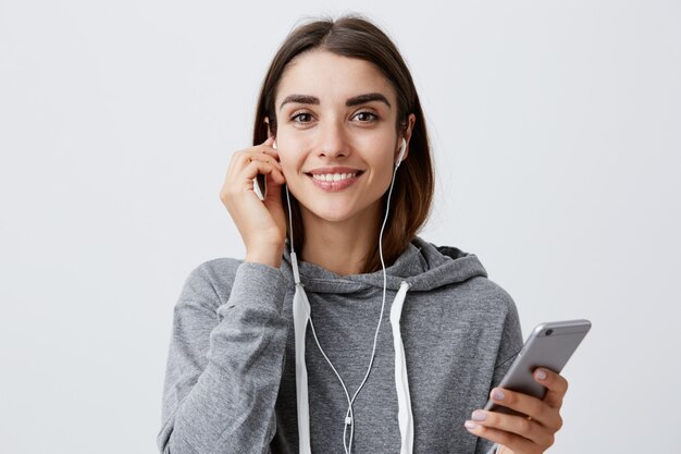 Mulher se preparando para um passeio no parque. Conceito de estilo de vida urbano. Jovem estudante morena bonito caucasiano com capuz cinza, sorrindo com dentes, usando fones de ouvido, segurando o smartphone nas mãos.