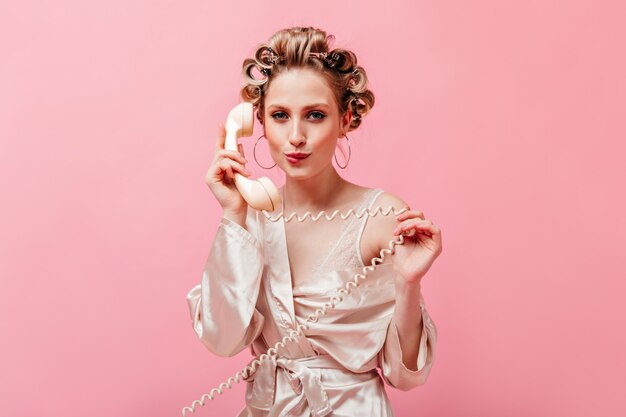 Foto grátis mulher sedutora com bobes de cabelo morde o lábio e fala no telefone fixo na parede rosa