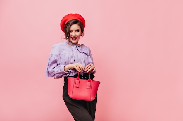 Mulher sedutora na boina, blusa e calças segurando uma bolsa vermelha em fundo rosa.