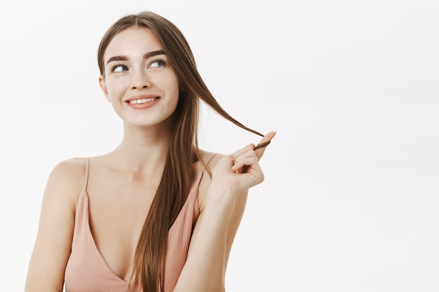 Mulher sedutora tendo pensamentos maliciosos em mente enrolando mecha de cabelo no dedo e sorrindo curiosamente com alguma intenção pensando ou sonhando olhando para o canto superior direito com um sorriso encantado