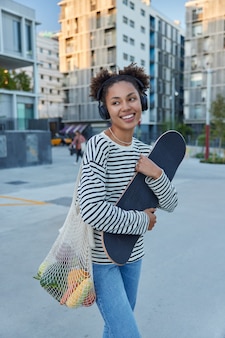 Mulher segura bolsa de lona carregada de skate com vegetais ouve música enquanto caminha pelas ruas da cidade vestida com um macacão listrado e jeans e aproveita o tempo de lazer