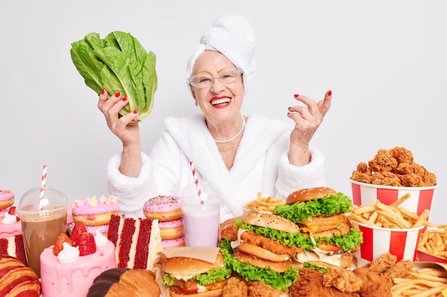 Foto grátis mulher segura vegetal verde prefere comer comida saudável em vez de refeição enganadora senta à mesa usa óculos transparentes, roupão de banho e toalha na cabeça cercada por produtos de alto teor calórico