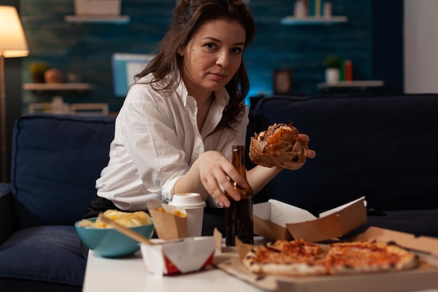 Mulher segurando a garrafa de cerveja, comendo saboroso hambúrguer para viagem enquanto relaxa no sofá em frente à mesa de tv com menu de fast food. pessoa com fome, desfrutando de junk food, sentado no sofá à noite.