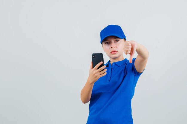 Mulher segurando o celular com o polegar para baixo em camiseta azul e boné