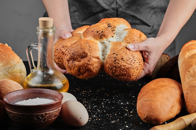 Mulher segurando pão na mesa escura com ovos, tigela de farinha e copo de óleo.