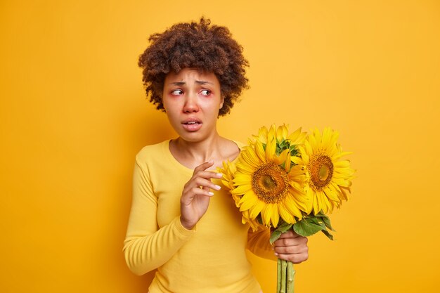 mulher segurando um buquê de girassóis tem olhos vermelhos e usa um macacão casual isolado sobre amarelo vivo