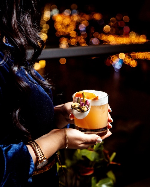 Foto grátis mulher segurando um copo de cocktail laranja, olhando a vista da cidade à noite