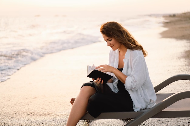 Mulher segurando um livro nas mãos lendo o livro em um mar durante o pôr do sol