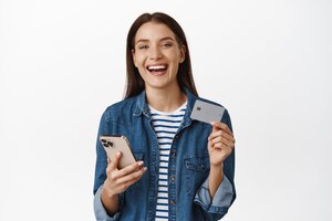 Mulher segurando um smartphone, mostrando o cartão de crédito, sorrindo satisfeita e rindo comprando smth no celular no branco
