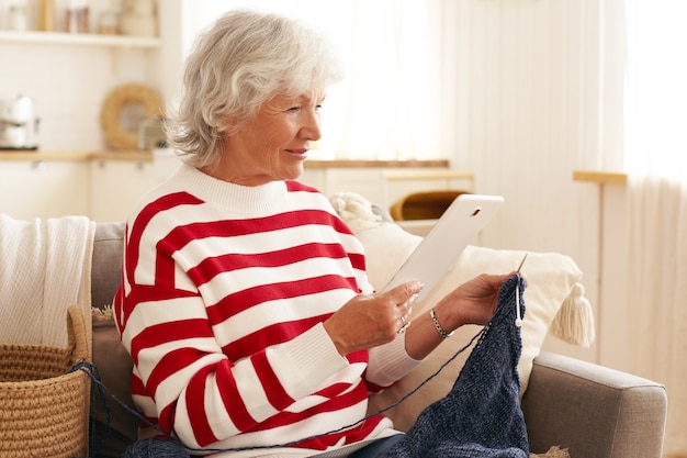 Mulher sênior de 60 anos com cabelos grisalhos, usando tablet digital dentro de casa. mulher idosa passando o tempo de lazer em casa, sentada no sofá, assistindo séries on-line em dispositivo eletrônico e tricotando