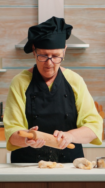 Mulher sênior desenrolando a massa para preparar pão caseiro. feliz chef idoso com bonete preparando ingredientes crus para assar pizza tradicional, polvilhando, peneirando a farinha na mesa da cozinha.