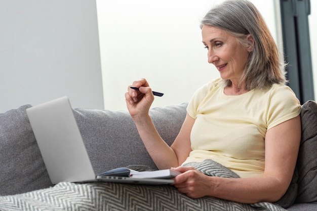 Mulher sênior em casa no sofá usando laptop e fazendo anotações