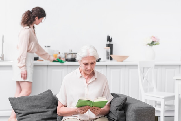 Mulher sênior, livro leitura, frente, mulher, fazendo, tarefas domésticas, trabalho