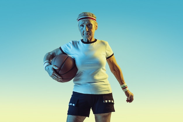 Mulher sênior no sportwear jogando basquete em fundo gradiente, luz de néon. Modelo feminino em ótima forma permanece ativo. Conceito de esporte, atividade, movimento, bem-estar, confiança. Copyspace.