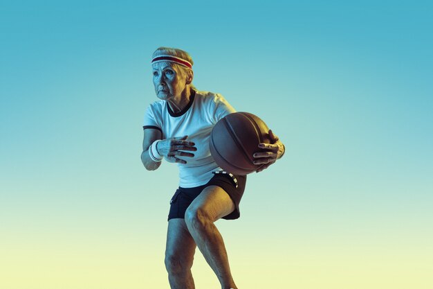 Mulher sênior no sportwear jogando basquete em fundo gradiente, luz de néon. Modelo feminino em ótima forma permanece ativo. Conceito de esporte, atividade, movimento, bem-estar, confiança. Copyspace.