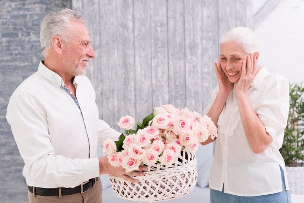 Mulher sênior surpreendida olhando para cesta de rosas, realizada por seu marido feliz