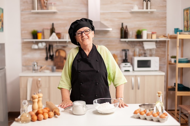 Mulher sênior vestindo uniforme de padeiro na cozinha de casa, sorrindo para a câmera. padeiro idoso aposentado em uniforme de cozinha, preparando ingredientes de pastelaria na mesa de madeira, pronta para cozinhar pão saboroso caseiro.