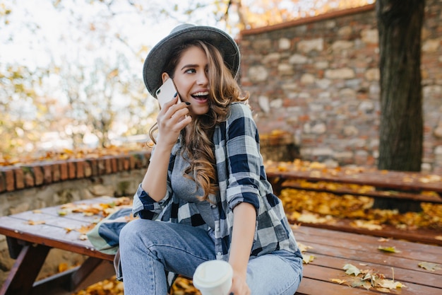 Mulher sensual e cacheada com chapéu expressando emoções engraçadas durante a sessão de fotos no jardim de outono