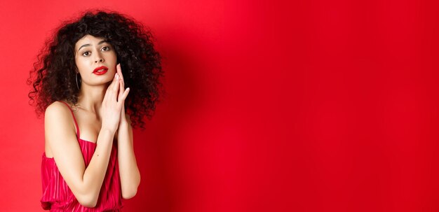 Foto grátis mulher sensual romântica com cabelo encaracolado usando vestido de noite posando sedutora em fundo vermelho