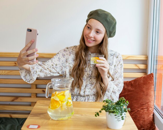 Mulher sentada à mesa com um tiro no meio, segurando um copo de limonada e tirando uma selfie