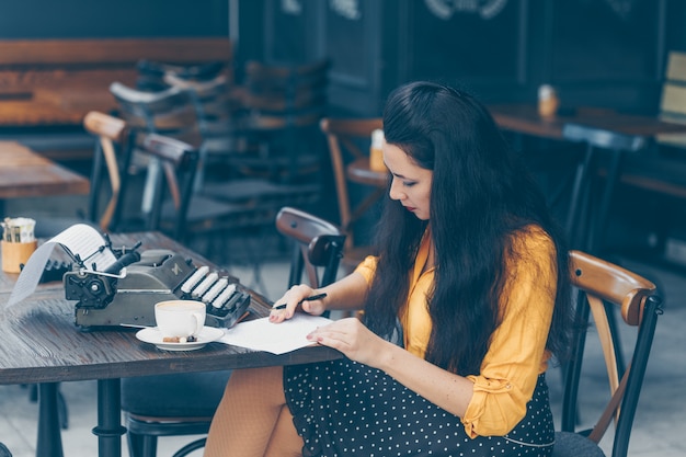 Mulher sentada e escrevendo algo no papel e pensativo e em amarelo top e saia longa no terraço do café durante o dia.