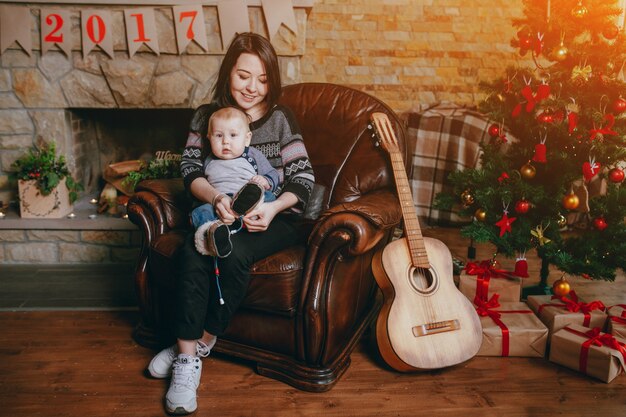 Mulher sentada em uma única poltrona com seu bebê e uma guitarra ao lado dele