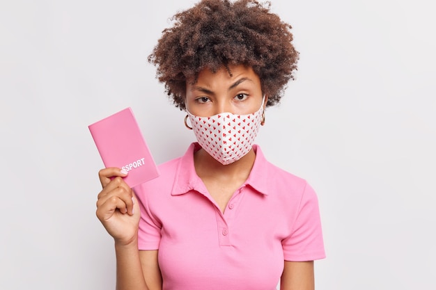 Foto grátis mulher séria com cabelo encaracolado com passaporte que vai viajar usa máscara protetora como proteção contra coronavírus evita infecção isolada sobre parede branca