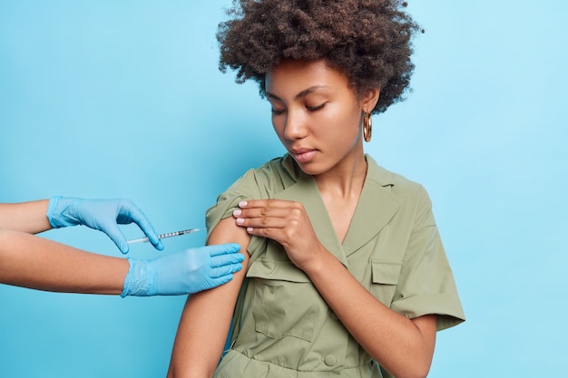 Mulher séria com cabelo encaracolado recebe vacina injetável contra a doença do coronavírus usa vestido verde isolado sobre a parede azul