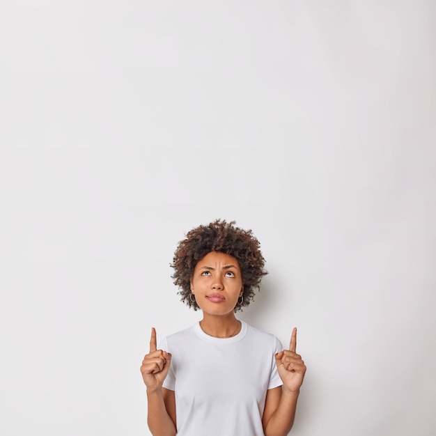 Foto grátis mulher séria de cabelos encaracolados focada atentamente acima indica os dedos indicadores para cima mostra propaganda ou promoção vestida casualmente demonstra espaço em branco para o seu conteúdo publicitário