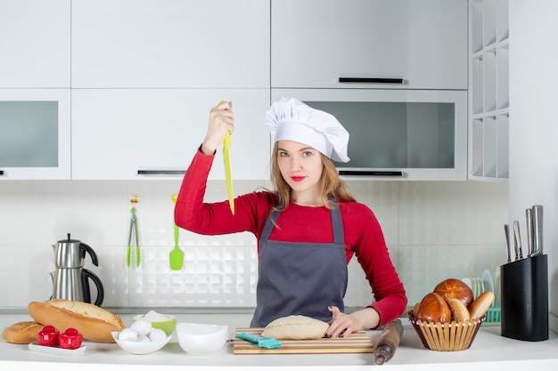 Mulher séria de frente com chapéu de cozinheira segurando uma faca na cozinha