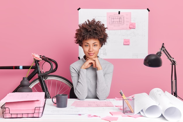 Foto grátis mulher séria, de pele escura, prepara projeto de construção ou poses de designer de interiores na área de trabalho com projetos de papel no escritório