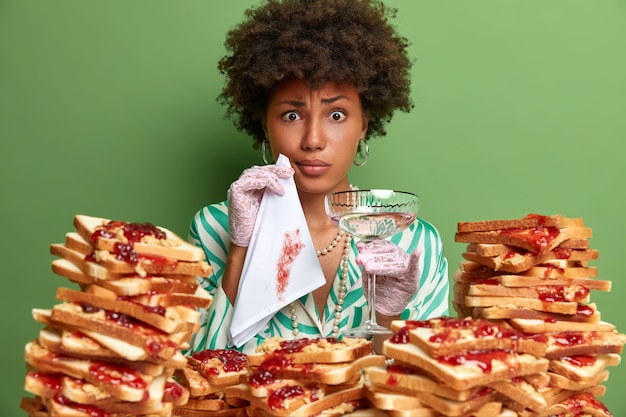 Mulher séria e descontente parece estranha, limpa a boca suja com guardanapo, vestida elegantemente, segura um copo de bebida alcoólica, posa perto de uma pilha de pão, isolada na parede verde. senhora no restaurante