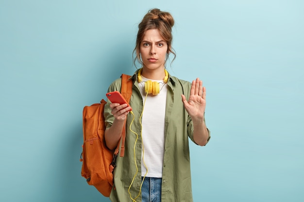 Mulher séria e fofa passa o tempo dentro de casa com um gadget moderno, mostra gesto de parar, pede para ir mais devagar, viaja com música e mochila